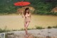 A naked woman holding a red umbrella by a lake.